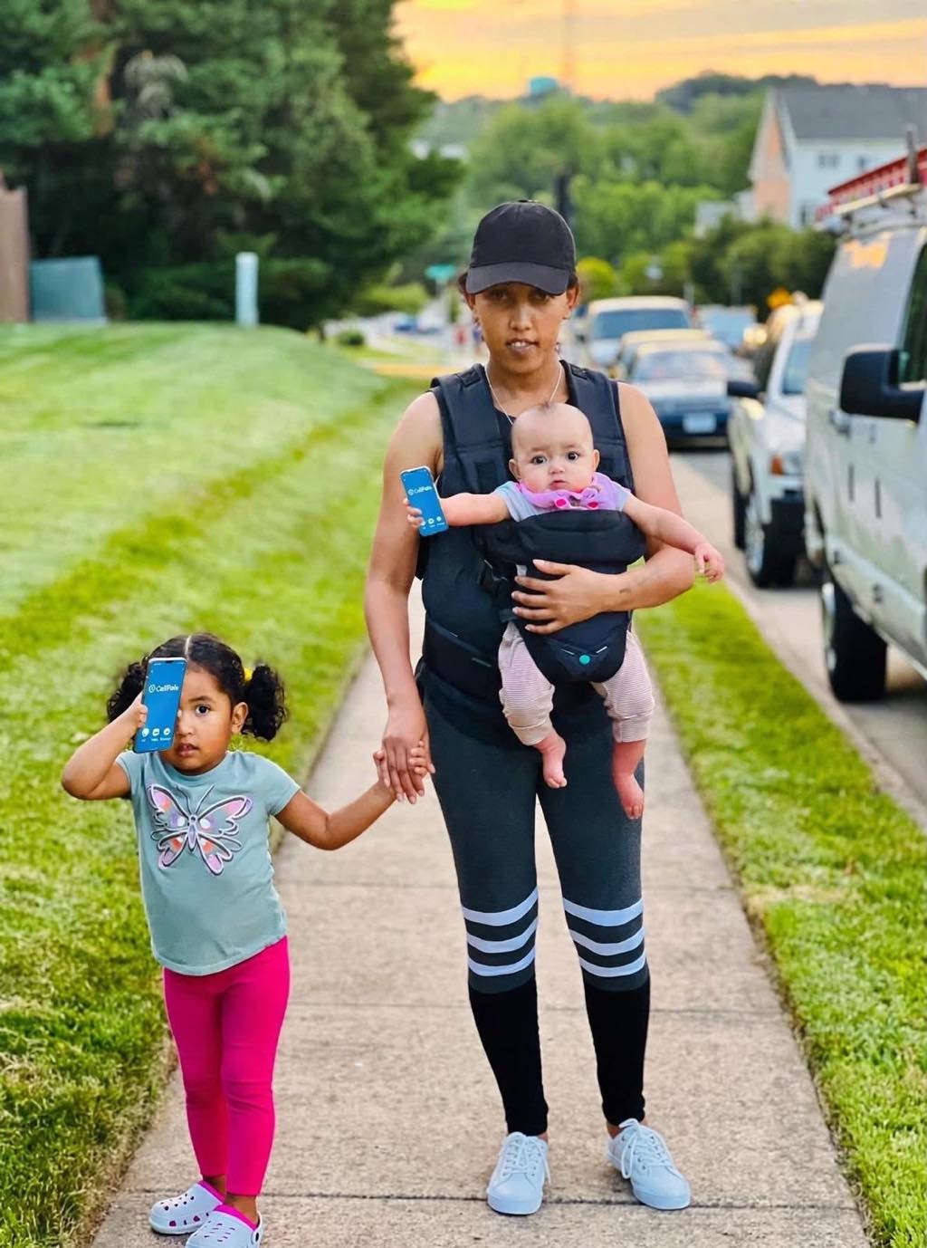 Mother walking at sunset holding hands with her toddler daughter and carrying her baby, both kids holding blue CallPalo phones
